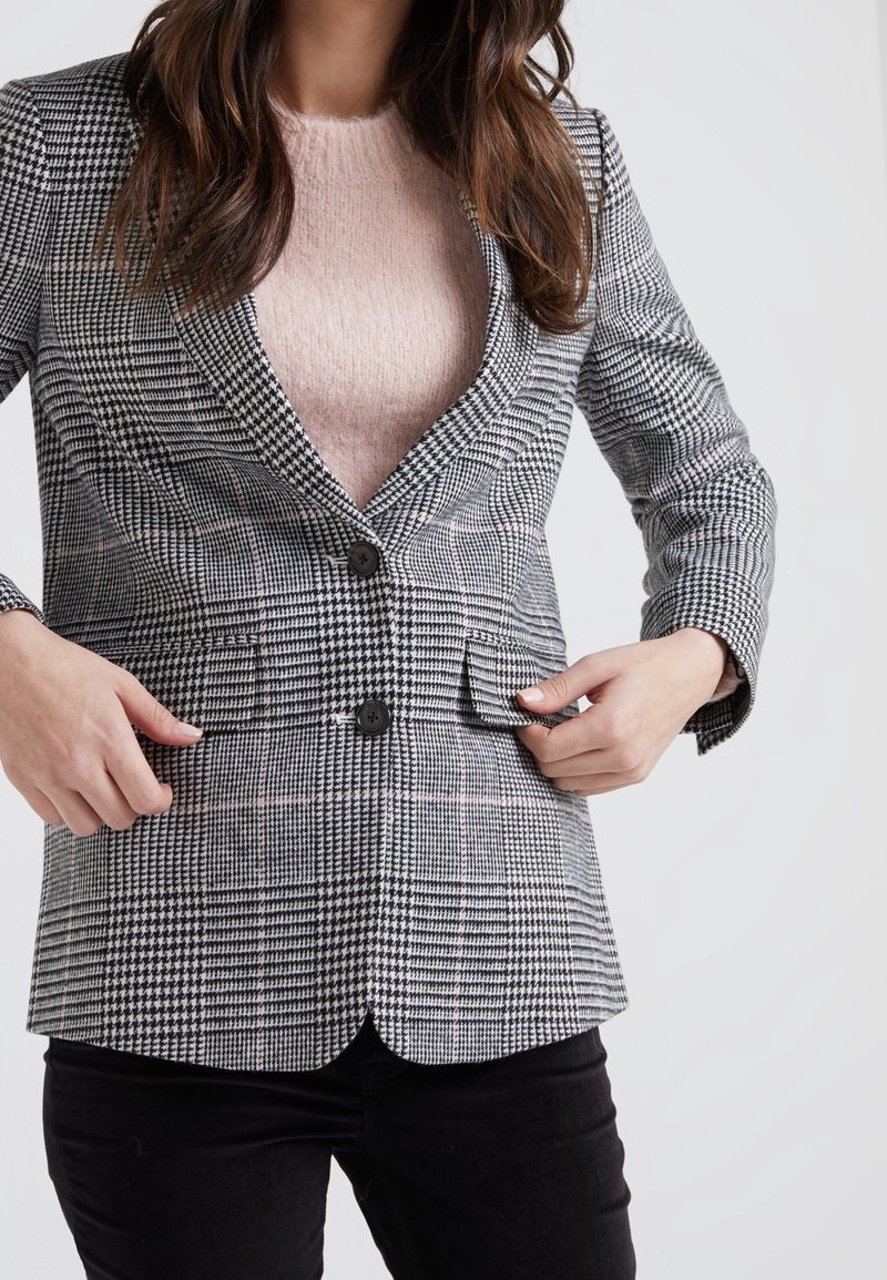 Femme ajustant un blazer à carreaux noir et blanc porté sur un pull duveteux rose clair et associé à un pantalon noir, sur un fond clair.