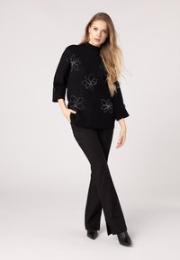Pullover nero oversize con ricami floreali, maniche a tre quarti e due bottoni ai polsini. Abbinato a pantaloni neri a zampa e stivaletti a punta.