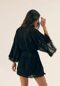 Przezroczysty czarny kimono z falbaniastymi rękawami, zwężoną talią i zwiewnym dołem. Lekka tkanina podkreśla delikatną teksturę i elegancki krój.