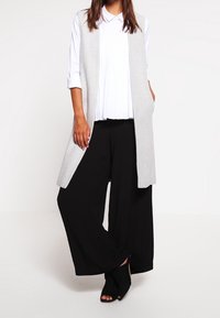 Grå ärmlös väst över en vit plisserad skjorta och svarta vida byxor, matchat med svarta sandaletter. Slät tyg, avslappnad passform.