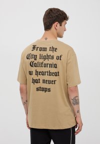 Beżowy, oversize t-shirt z wyrazistym, czarnym tekstem na plecach. Wykonany z bawełny, ma luźny krój i krótkie rękawy.