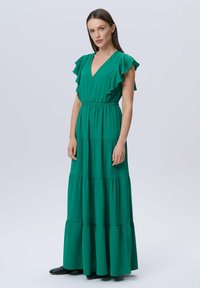 Vestido maxi verde con escote en V, mangas cortas con volantes, cinturilla elástica y falda en capas. Tela suave con una textura fluida.