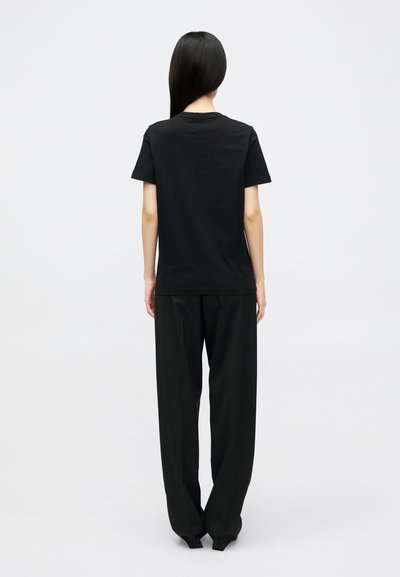 Άτομο με μακριά μαύρα μαλλιά φορώντας απλή μαύρη κοντομάνικη μπλούζα και χαλαρούς μαύρους παντελόνες, που στέκεται με πλάτη σε λευκό φόντο.