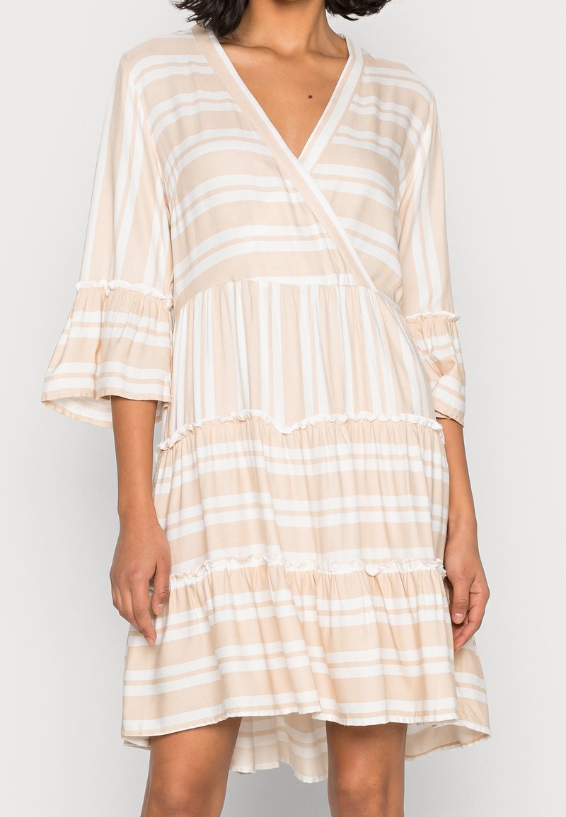 Femme portant une robe beige et blanche à rayures, col en V, avec des volants en étages et des manches cloche aux trois quarts, debout devant un fond uni.