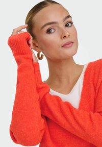 Femme aux cheveux plaqués en arrière portant un pull orange vif et des boucles d'oreilles créoles dorées, ajustant une manche près de son visage.