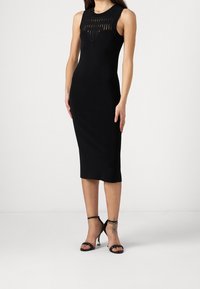 Abito midi nero con design senza maniche, caratterizzato da un sovrapposto testurizzato sul corpino e una silhouette aderente. Accentato da tacchi a strappo neri.