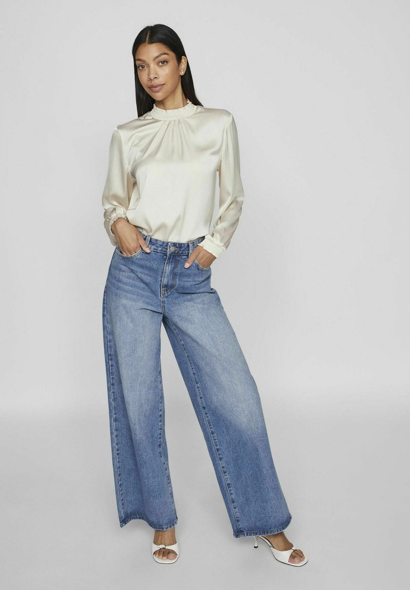 Blouse en satin crème avec des manches bouffantes et un col volanté, associée à un jean large taille haute bleu clair et des sandales à talons blanches.