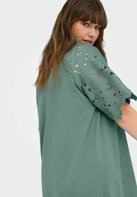 Tricou verde dintr-un material moale, cu detalii din dantelă florală pe mâneci, cu margini ondulate și modele decupate.