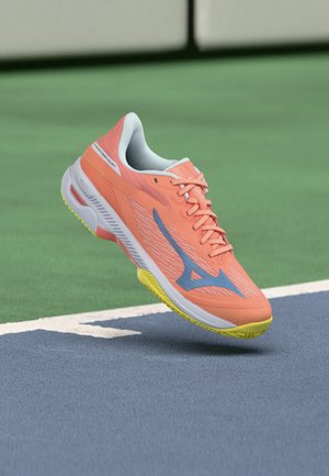 Chaussure de sport orange vif avec un logo bleu et une semelle jaune flottant au-dessus d’une surface de court de tennis bleu et vert près d’une ligne blanche.
