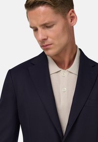 Tumed bleiser kergelt beeži polo särgi kohal, millel on kolm nuppu ja tasku. Bleiseril on peen disain ja sile tekstuur.