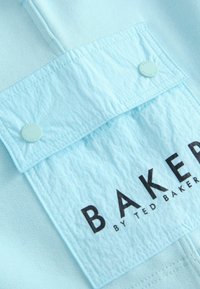 Tessuto blu chiaro con una tasca sul petto testurizzata, dotata di un bottone a pressione e un bold testo nero che recita "BAKED BY TED BAKER".