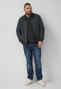 Ciemna, fakturowana kurtka na zamek nałożona na szary sweter, w połączeniu z niebieskimi dżinsami i jasnymi sneakersami. Zestaw charakteryzuje się prostymi liniami i neutralnymi odcieniami.