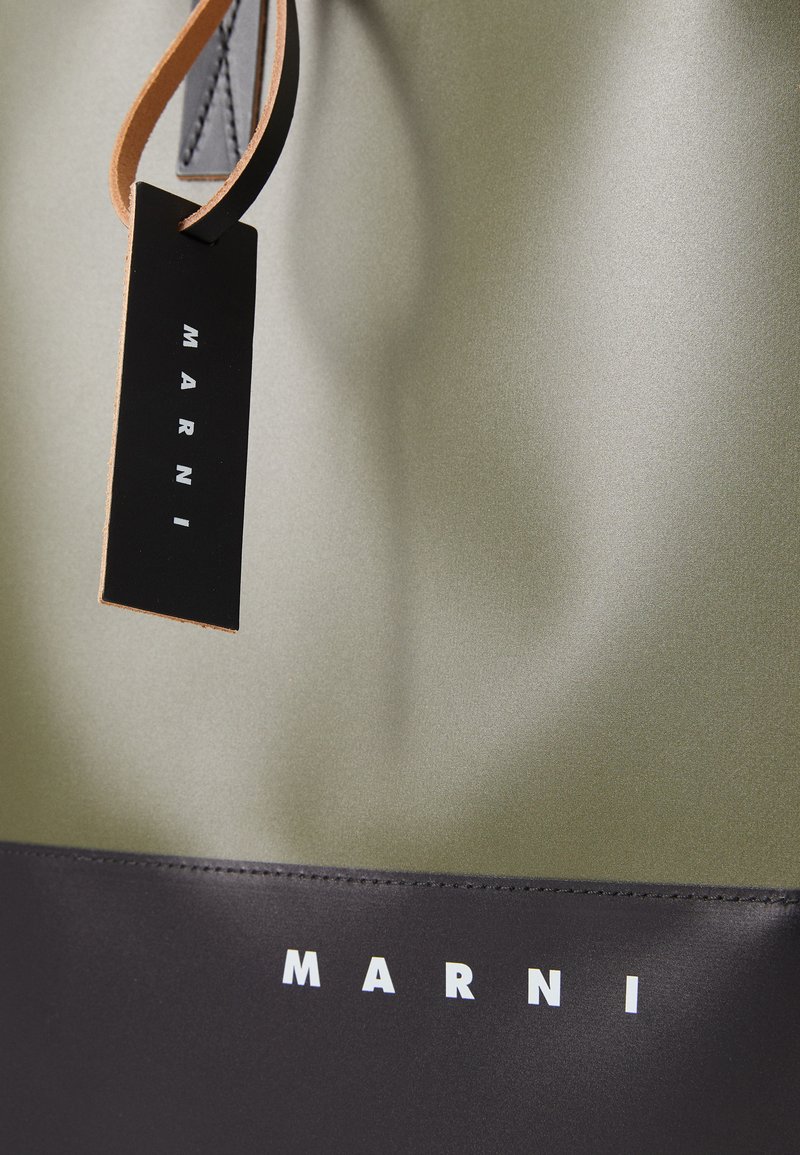 Borsa tote verde oliva con una texture liscia, fondo nero, testo del logo bianco e un'etichetta in pelle con una tracolla marrone.