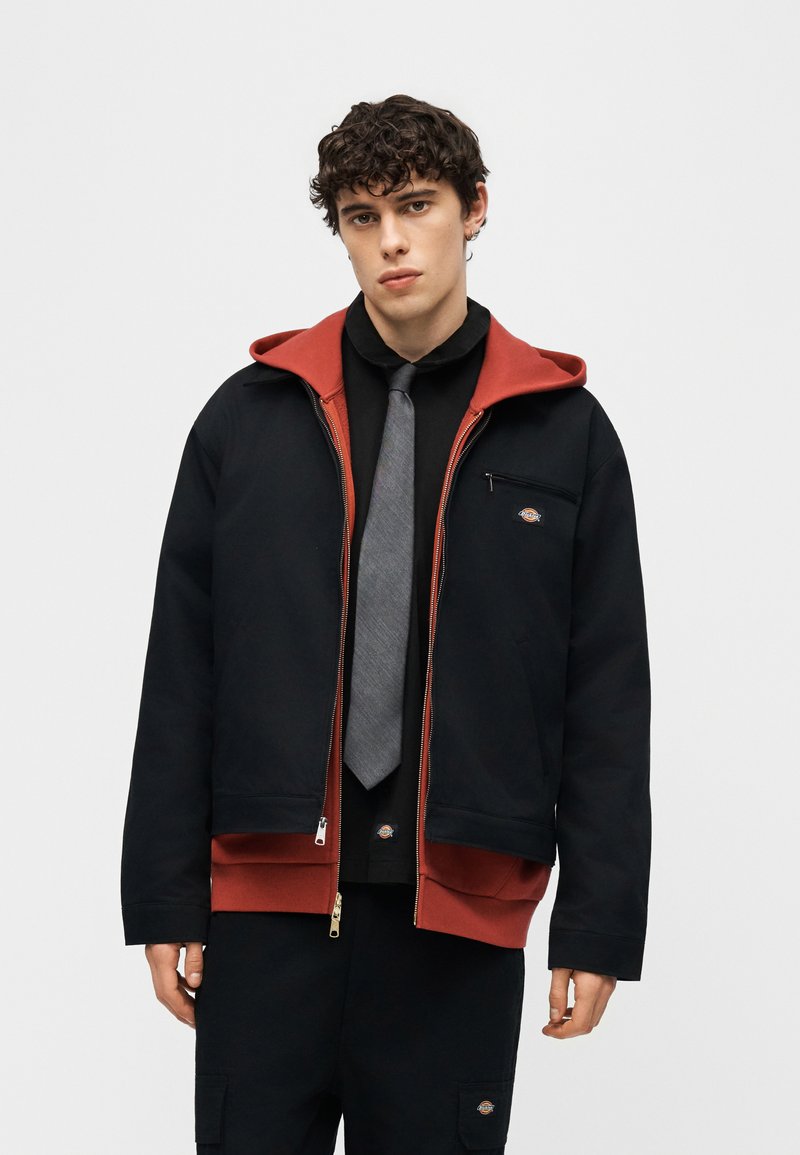 Junger Mann mit lockigem Haar, der eine schwarze Jacke über einem roten Hoodie, einem schwarzen Hemd, einer grauen Krawatte und einer schwarzen Hose vor einem einfarbigen Hintergrund trägt.