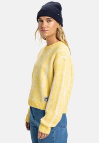 Geel gestreepte gebreide trui met een ribbelboord aan de ronde hals en brede manchetten. Gecombineerd met blauwe denim jeans en een donkere beanie.