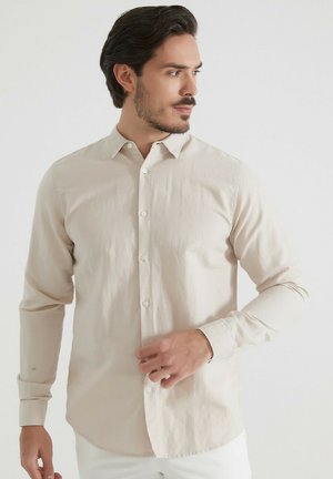 Homme aux cheveux foncés portant une chemise beige à manches longues boutonnée, regardant vers sa droite, ajustant le devant de la chemise.