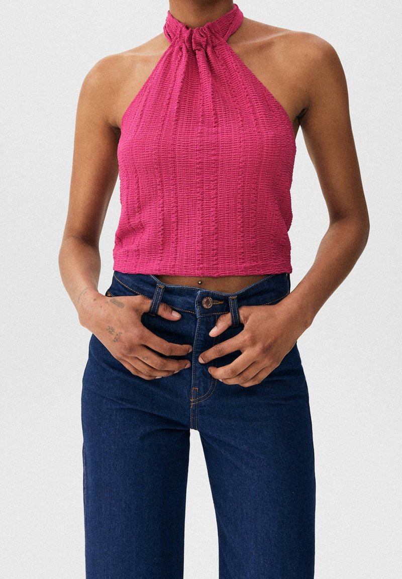 Femme portant un haut halter sans manches magenta avec un col froncé et un jean bleu foncé taille haute, mains ajustant la ceinture.