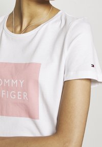 Camiseta de algodón blanca con un logotipo rectangular de color rosa claro que incluye el texto "TOMMY HILFIGER". Mangas cortas con detalle del logotipo en la manga.