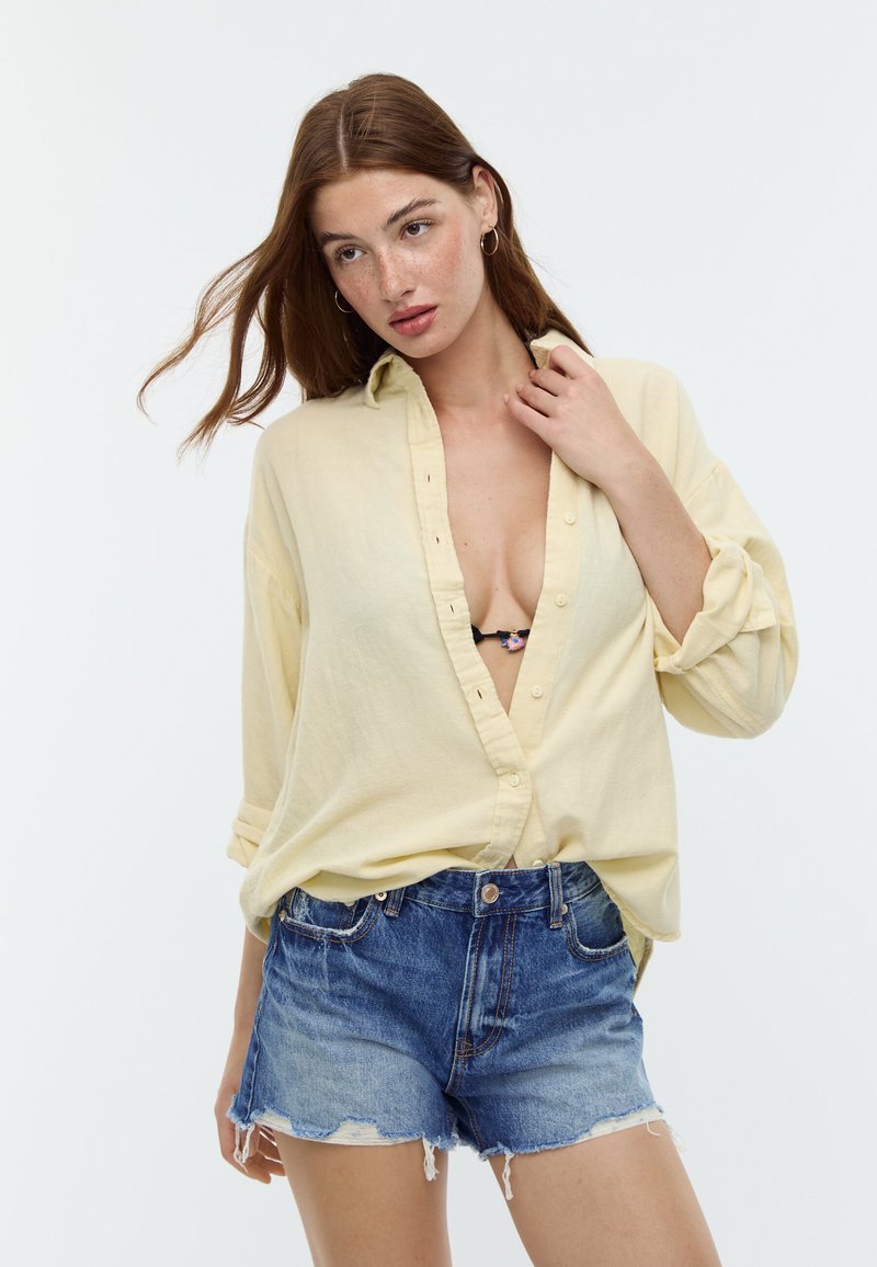 Ung kvinde med lysebrunt hår iført en lys gul skjorte med knapper over en sort bh og slidte blå denimshorts, poserer mod hvid baggrund.