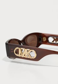 Ochelari de soare din acetat maro, cu detalii aurii ale logo-ului MK pe brațe, lentile rotunde și o finisare netedă și lucioasă. Prezentă un design cu decupaj pe ramă.