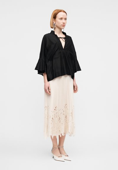 Μαύρο μεταξωτό μπλουζάκι με φαρδιές μανσέτες και δετή λαιμόκοψη, σε συνδυασμό με μια κρεμ πλισέ φούστα με κεντήματα λουλουδιών και λευκά τακούνια με λουράκι στον αστράγαλο.