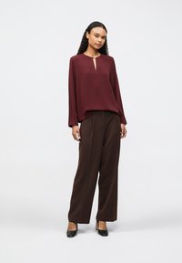 Μπορντό μπλούζα με λαιμόκοψη σε σχήμα κλειδαρότρυπας, συνδυασμένη με καφέ παντελόνι ψηλόμεσο, φαρδύ. Το μοντέλο φοράει μαύρες γόβες, με τα μαλλιά σε χαλαρούς κυματισμούς.