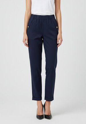 Donna che indossa pantaloni blu navy a sigaretta con orli risvoltati e dettagli di bottoni dorati sulle tasche, abbinati a scarpe con tacco a punta nere e una maglia bianca.