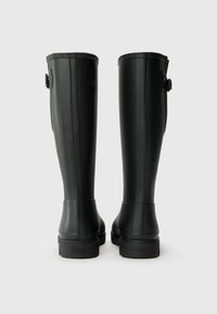 Bottes de pluie en caoutchouc noires avec une texture lisse, des rainures verticales et un détail de boucle ajustable en haut. Semelles robustes avec un léger talon.