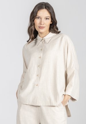 Kobieta z falującymi brązowymi włosami nosi luźną, jasnobeżową koszulę zapinaną na guziki oraz pasujące spodnie, z rękami w kieszeniach na jednolitym tle.