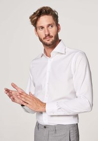 Chemise blanche à manches longues avec un col classique, fermeture à bouton et texture de tissu lisse, portée avec un pantalon gris. Mains positionnées devant.