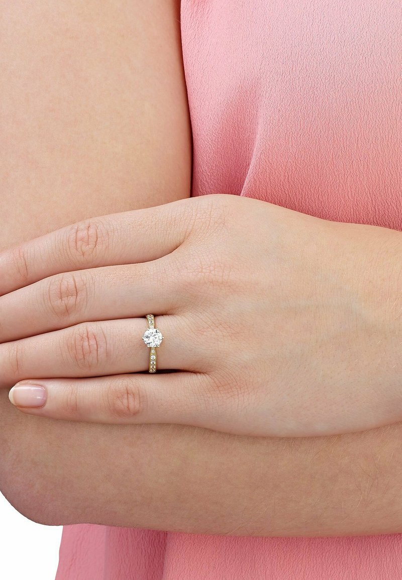 Gouden verlovingsring met een ronde, fonkelende diamant in het midden, verfraaid met kleine, heldere stenen langs de band. Hand in huidskleur die rust.