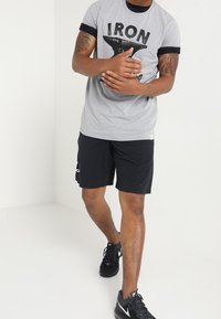 Harmaa T-paita mustilla yksityiskohdilla ja "IRON"-grafiikalla, yhdistettynä mustiin shortseihin, joissa on sivutaskut ja valkoinen logo. Mustat urheilukengät.