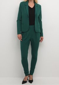 Blazer verde con pantalones tapered a juego, con una textura suave. Debajo lleva una blusa negra de escote en V. La modelo lleva tacones negros.