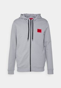 Sudadera gris con cremallera, con un bolsillo frontal, cordones ajustables negros y un parche con logo rojo en el pecho; textura de tela suave.