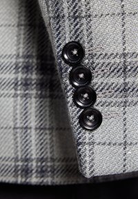 Jasnoszary wełniany żakiet w wzór houndstooth, ozdobiony czterema czarnymi guzikami na mankietach i subtelnymi niebieskimi akcentami.
