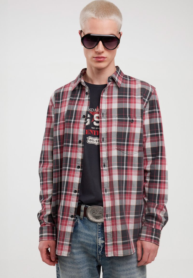 Rutig skjorte i rød, hvid og sort, med knaplukning, lange ærmer og en enkelt brystlomme, parret med blå denim jeans og solbriller.