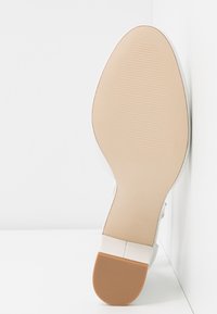Semelle d'une chaussure blanche à talon avec un dessous beige lisse et une pointe de talon marron texturée, positionnée sur fond blanc.