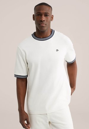 Moški, ki nosi belo srajco s kratkimi rokavi z mornarsko modrimi črtastimi ovratnikom in manšetami ter majhno vezeno logotipom na prsih, stoji ob nevtralni podlagi.