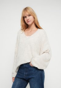 Suéter oversized bege claro com decote em V, punhos canelados e acabamento macio e texturizado, combinado com jeans azul escuro.