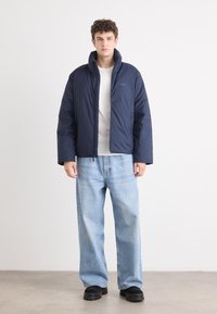 Navy pufferjack met hoge kraag, ritssluiting en een gladde textuur. Gecombineerd met lichtblauwe, losse jeans en zwarte schoenen.