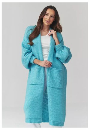 Kobieta z długimi brązowymi włosami, ubrana w długi, gruby, jasnoniebieski sweter na drutach, noszony na białej bluzce i białych spodniach.