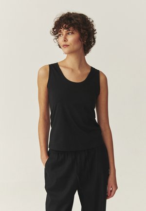 Jeune femme aux cheveux bouclés portant un haut noir sans manches et un pantalon noir, debout avec une main dans la poche, regardant sur le côté.