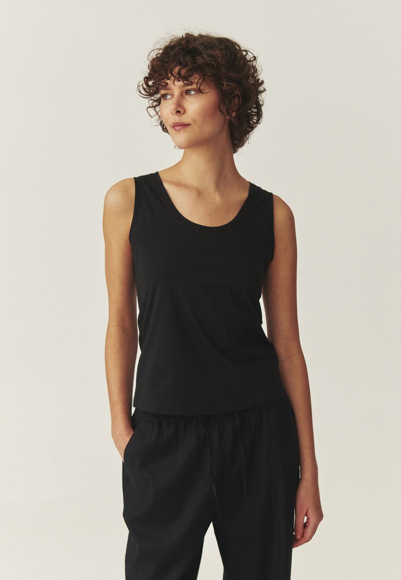 Jeune femme aux cheveux bouclés portant un haut noir sans manches et un pantalon noir, debout avec une main dans la poche, regardant sur le côté.