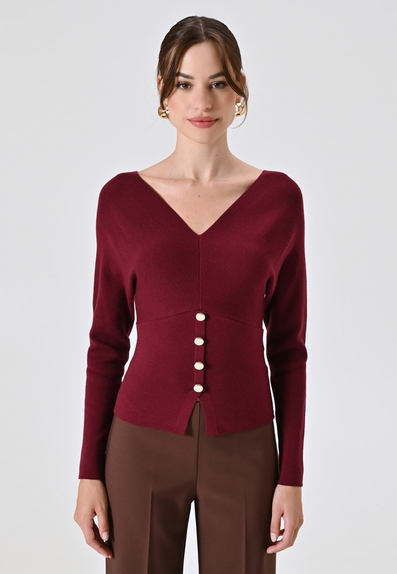 Maglione a maglia color bordeaux con scollo a V, caratterizzato da una fila di sei bottoni bianchi sul davanti e uno spacco all'orlo. Maniche lunghe.