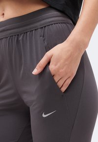 Χέρι που στηρίζεται στην τσέπη σκούρου γκρι αθλητικού παντελονιού Nike με ελαστική μέση, εμφανίζοντας την κοιλιακή περιοχή και τη μαύρη πάνω άκρη.