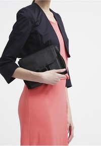 Svart läderklutch hålls under armen, med ett vikbart lock och minimal hårdvara, tillsammans med en korallfärgad klänning och en marinblå blazer.