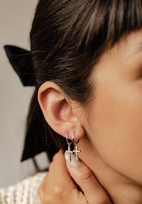 Boucles d'oreilles en argent avec des accents en cristal, en forme de croix, pendent d'une oreille percée. Cheveux coiffés en arrière avec un ruban noir.