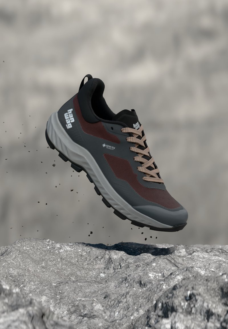 Παντόφλα πεζοπορίας σκούρου γκρι και μαρόν με μπεζ κορδόνια, ετικέτα Gore-Tex, αιωρείται πάνω από βραχώδες έδαφος με πτώση χαλαρού χαλικιού.