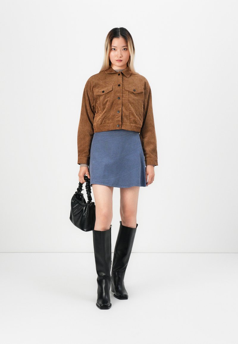 Femme portant une veste en velours côtelé marron, une jupe bleue, des bottes noires jusqu'au genou, tenant un sac à main noir, se tenant contre un fond blanc uni.