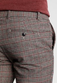 Szare spodnie w kratę z wzorem houndstooth, z czerwonymi akcentami, zapinanym na guzik tylnym kieszeniem i fakturowanym materiałem.